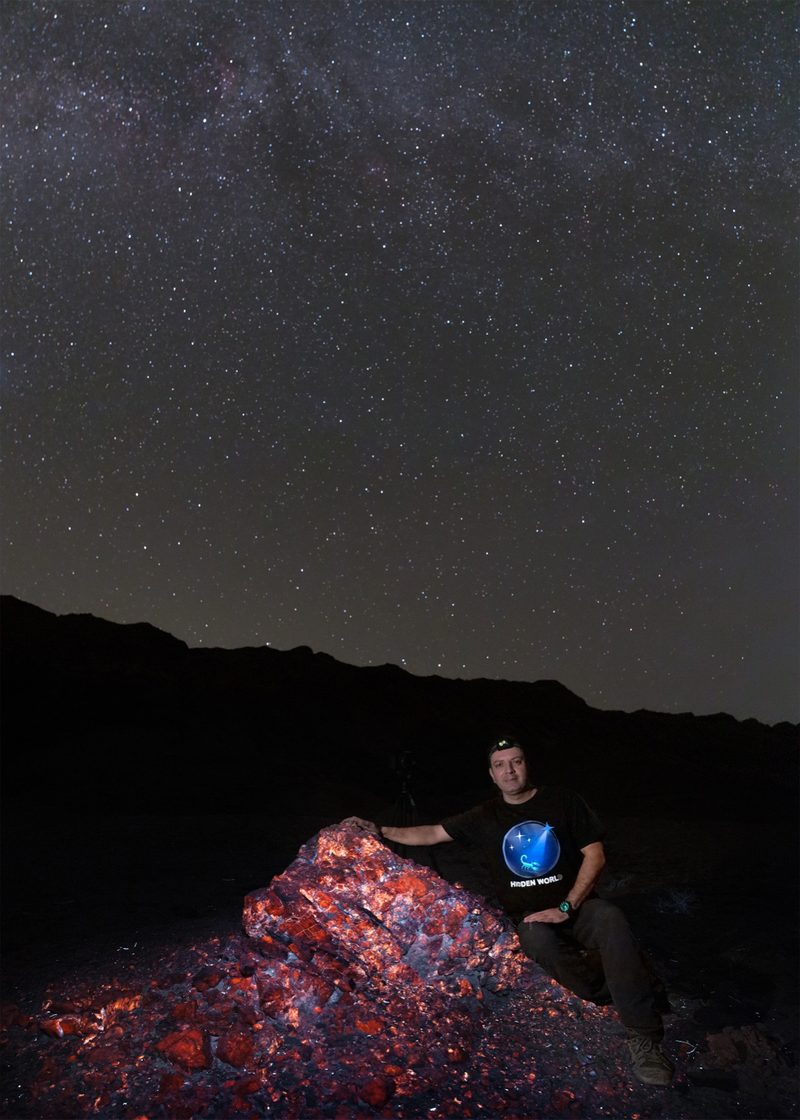 A photographer kneeling beside a large outcrop glowing orange under ultraviolet light, with the Milky Way overhead.