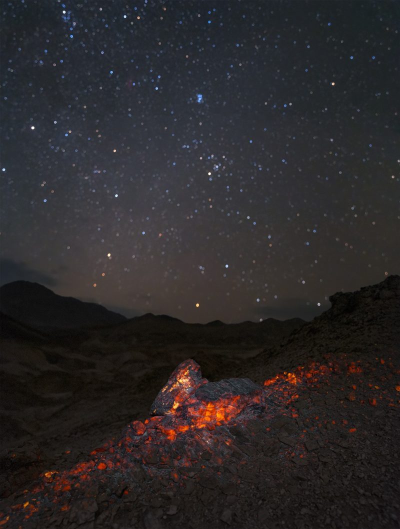 Desert floor strewn with rocks glowing orange under ultraviolet light, beneath a sky full of stars.